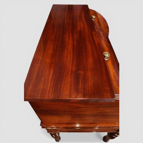 fig 10. An early nineteenth-century mahogany cylinder desk featuring a curved roll-front cover, brass knobs, a long frieze drawer, and turned reeded legs with brass castors.