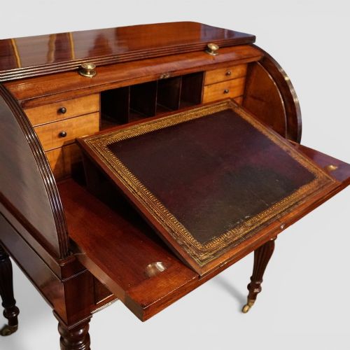 fig 11. An early nineteenth-century mahogany cylinder desk featuring a curved roll-front cover, brass knobs, a long frieze drawer, and turned reeded legs with brass castors.