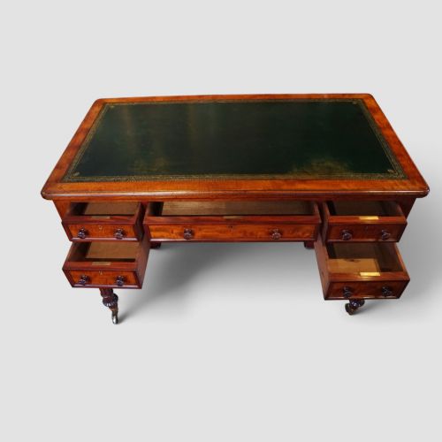 fig 3. Victorian mahogany writing table featuring a dark green leather top, five drawers with circular wooden handles, turned legs, and brass castors.