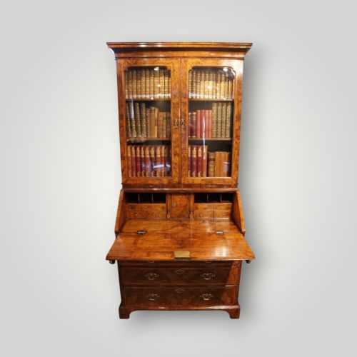 fig 1. A George I walnut bureau bookcase featuring glazed upper doors housing books, a slant-front writing flap, and lower drawers with brass drop handles.