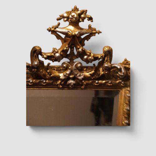 fig 2. An antique trapezoidal wall mirror featuring an ornate giltwood frame with silver-gilt accents, a decorative carved crest, and a tapered profile.