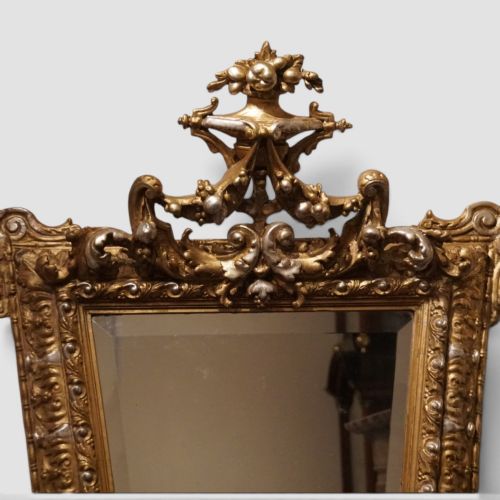 fig 3. An antique trapezoidal wall mirror featuring an ornate giltwood frame with silver-gilt accents, a decorative carved crest, and a tapered profile.