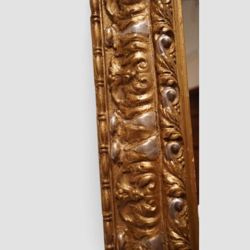fig 4. An antique trapezoidal wall mirror featuring an ornate giltwood frame with silver-gilt accents, a decorative carved crest, and a tapered profile.