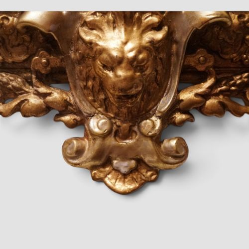 fig 6. An antique trapezoidal wall mirror featuring an ornate giltwood frame with silver-gilt accents, a decorative carved crest, and a tapered profile.