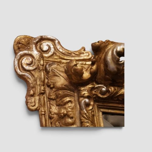 fig 8. An antique trapezoidal wall mirror featuring an ornate giltwood frame with silver-gilt accents, a decorative carved crest, and a tapered profile.