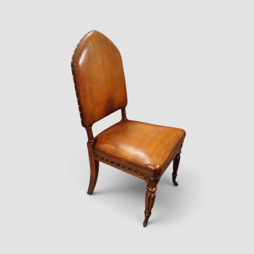 fig 1. A set of four Victorian oak dining chairs featuring arched gothic-style backrests upholstered in warm brown leather with brass stud trim.