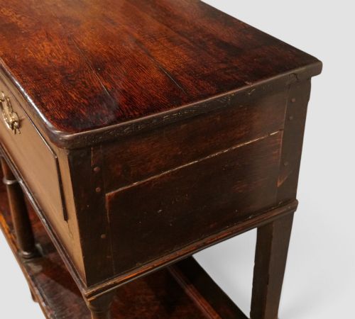 fig 3. An antique dark oak dresser base featuring three frieze drawers with brass handles supported by turned legs and a low open potboard shelf.