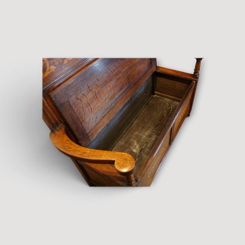 fig 6. An Art Nouveau solid oak settle featuring decorative marquetry inlay across the backrest and lower front panels, curved armrests, and a lift-up storage seat.