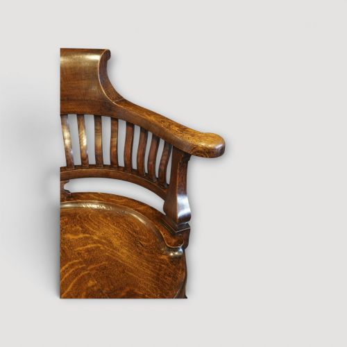 fig 2. An Edwardian dark oak revolving desk chair featuring a curved slatted backrest, shaped wooden saddle seat, and four legs with castors.
