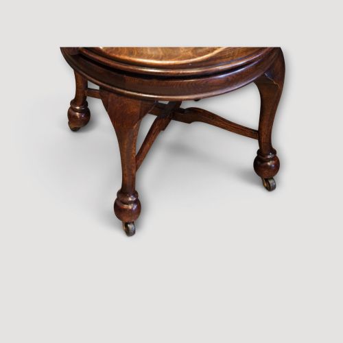 fig 3. An Edwardian dark oak revolving desk chair featuring a curved slatted backrest, shaped wooden saddle seat, and four legs with castors.