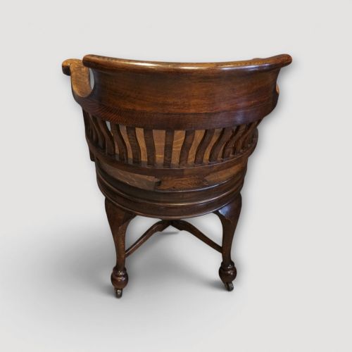 fig 6. An Edwardian dark oak revolving desk chair featuring a curved slatted backrest, shaped wooden saddle seat, and four legs with castors.