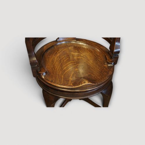 fig 7. An Edwardian dark oak revolving desk chair featuring a curved slatted backrest, shaped wooden saddle seat, and four legs with castors.