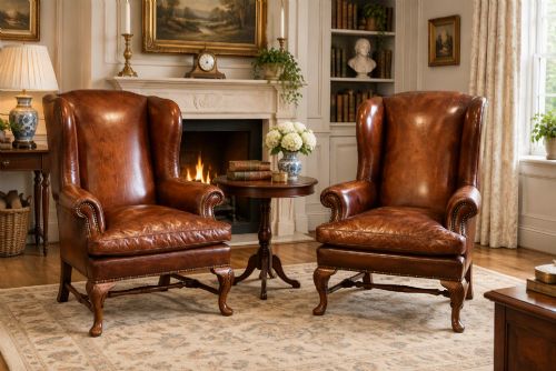 fig 6. A pair of brown leather Queen Anne style wingback armchairs featuring deep seat cushions, brass stud detailing, and carved walnut cabriole legs.