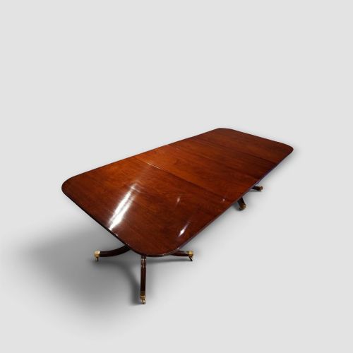 fig 1. An extended mahogany twin pedestal dining table featuring a polished surface, rounded corners, reeded tripod legs, and brass castors.