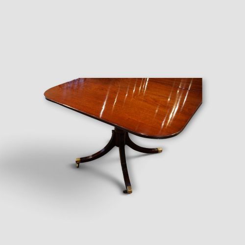 fig 6. An extended mahogany twin pedestal dining table featuring a polished surface, rounded corners, reeded tripod legs, and brass castors.