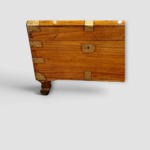 fig 3. A low rectangular warm amber camphor wood campaign trunk featuring brass strap corners, flush brass handles, dual keyholes, and turned legs with castors.