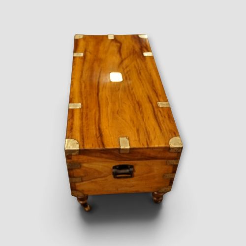 fig 4. A low rectangular warm amber camphor wood campaign trunk featuring brass strap corners, flush brass handles, dual keyholes, and turned legs with castors.