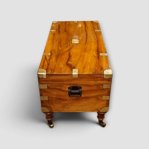 fig 5. A low rectangular warm amber camphor wood campaign trunk featuring brass strap corners, flush brass handles, dual keyholes, and turned legs with castors.