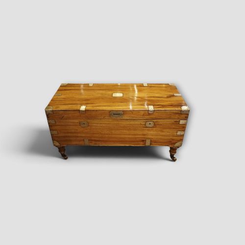 fig 6. A low rectangular warm amber camphor wood campaign trunk featuring brass strap corners, flush brass handles, dual keyholes, and turned legs with castors.