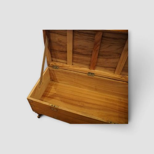 fig 8. A low rectangular warm amber camphor wood campaign trunk featuring brass strap corners, flush brass handles, dual keyholes, and turned legs with castors.