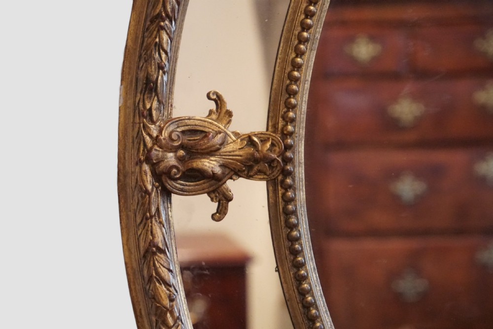 An antique oval wall mirror featuring an ornate gilded frame adorned with a ribbon and wreath crest, leaf motifs, and a beaded inner border. View VI