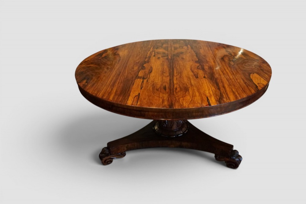 A circular William IV rosewood dining table with a figured veneer top on a central pedestal and tripartite base with scroll feet.