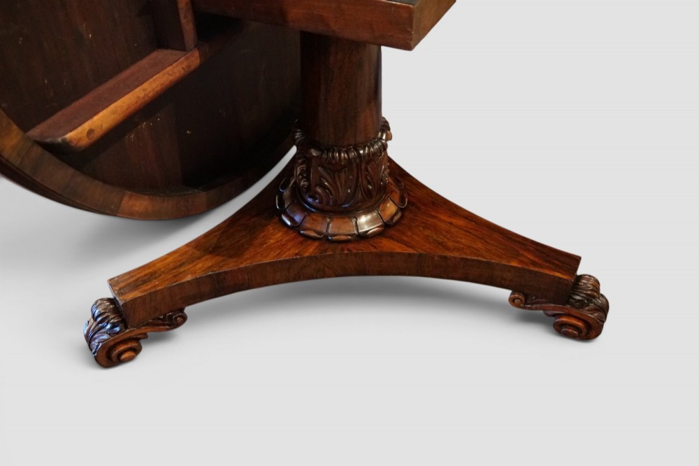 A circular William IV rosewood dining table with a figured veneer top on a central pedestal and tripartite base with scroll feet. View II