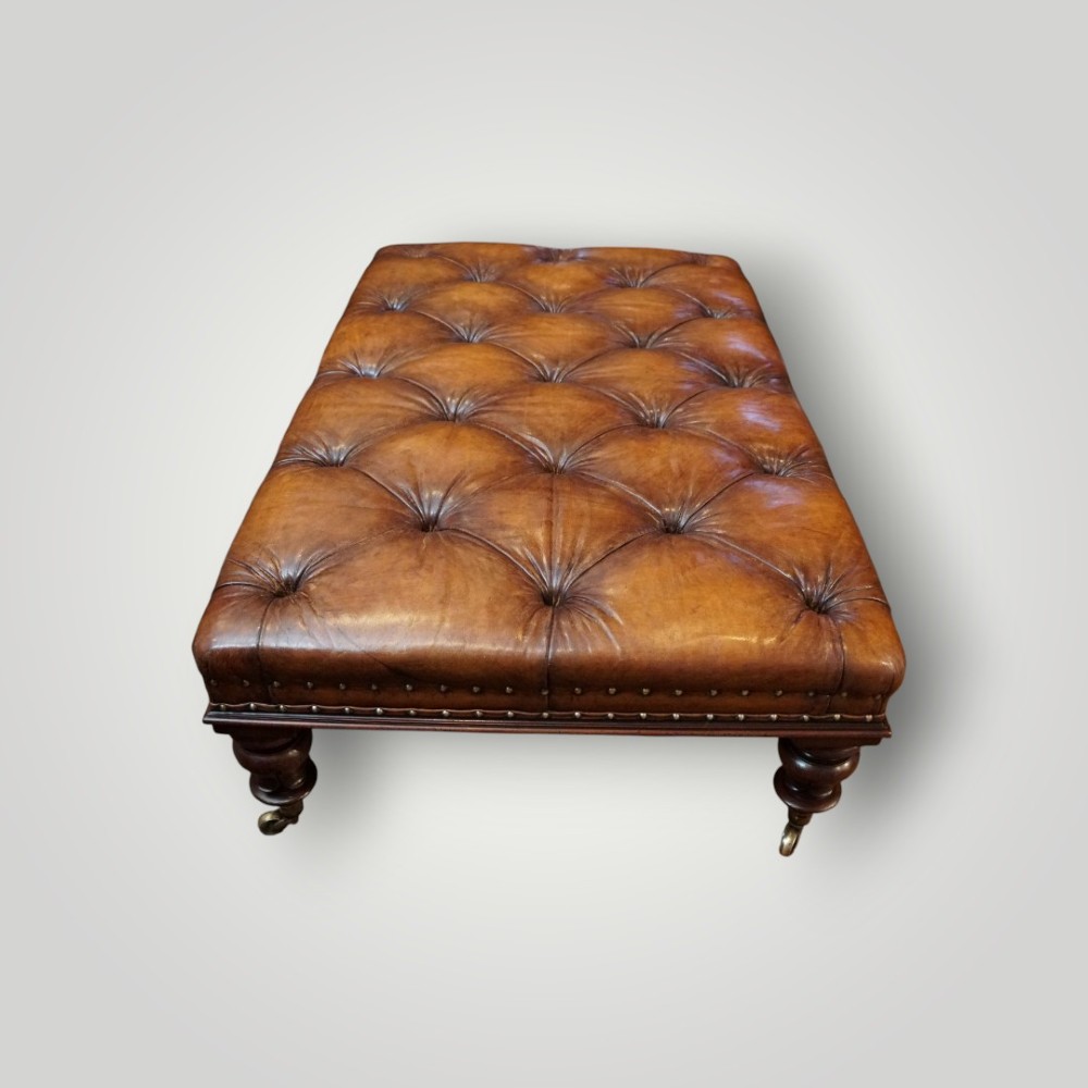 A large rectangular deep-buttoned tan leather Chesterfield footstool featuring brass stud trim, turned timber legs, and brass castors. View V