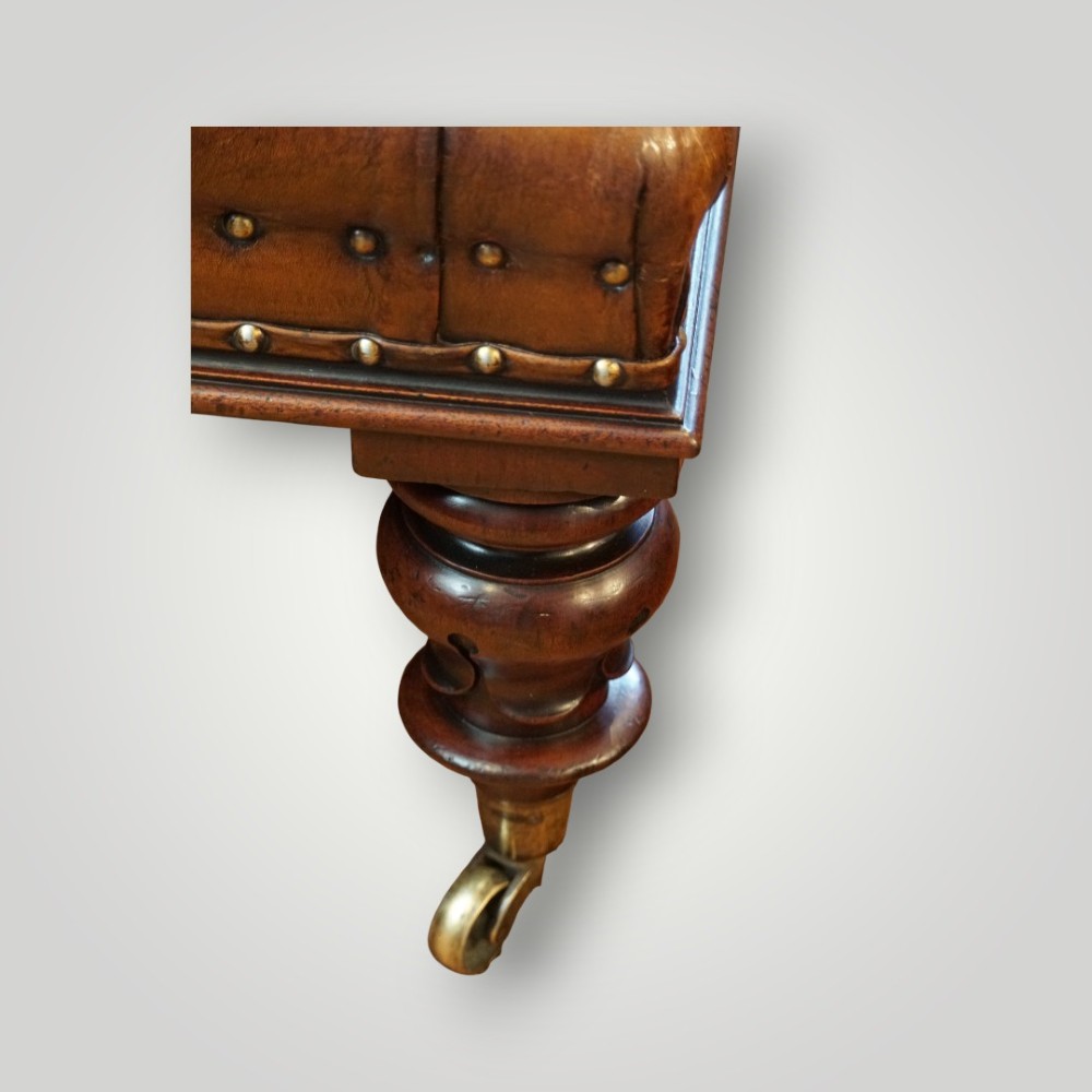 A large rectangular deep-buttoned tan leather Chesterfield footstool featuring brass stud trim, turned timber legs, and brass castors. View VI