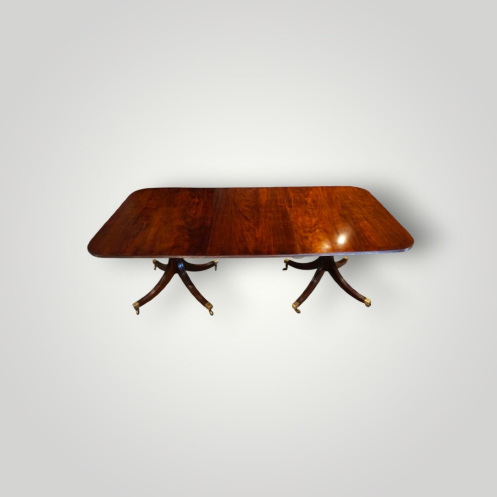 A polished mahogany twin pillar dining table featuring rounded corners, two turned central pedestals, and splayed legs resting on brass castors.