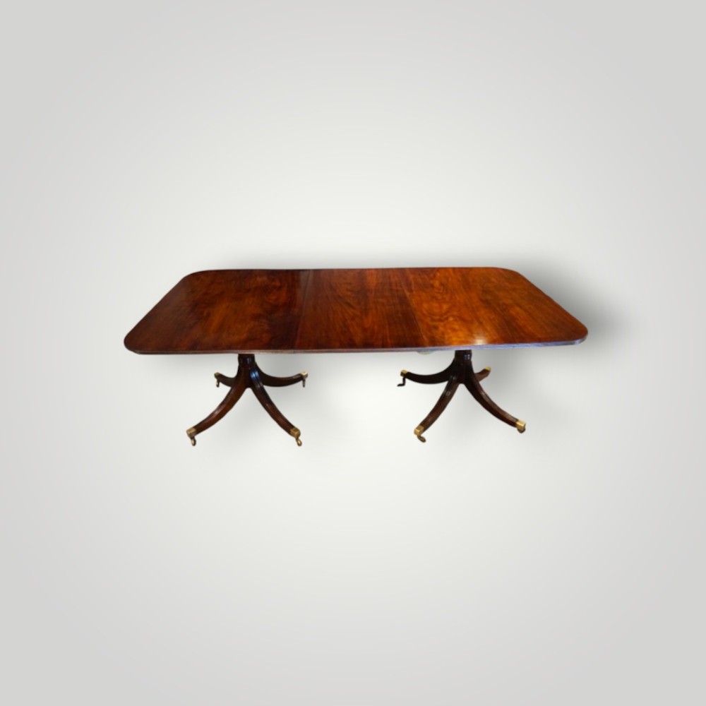 A polished mahogany twin pillar dining table featuring rounded corners, two turned central pedestals, and splayed legs resting on brass castors. View VII