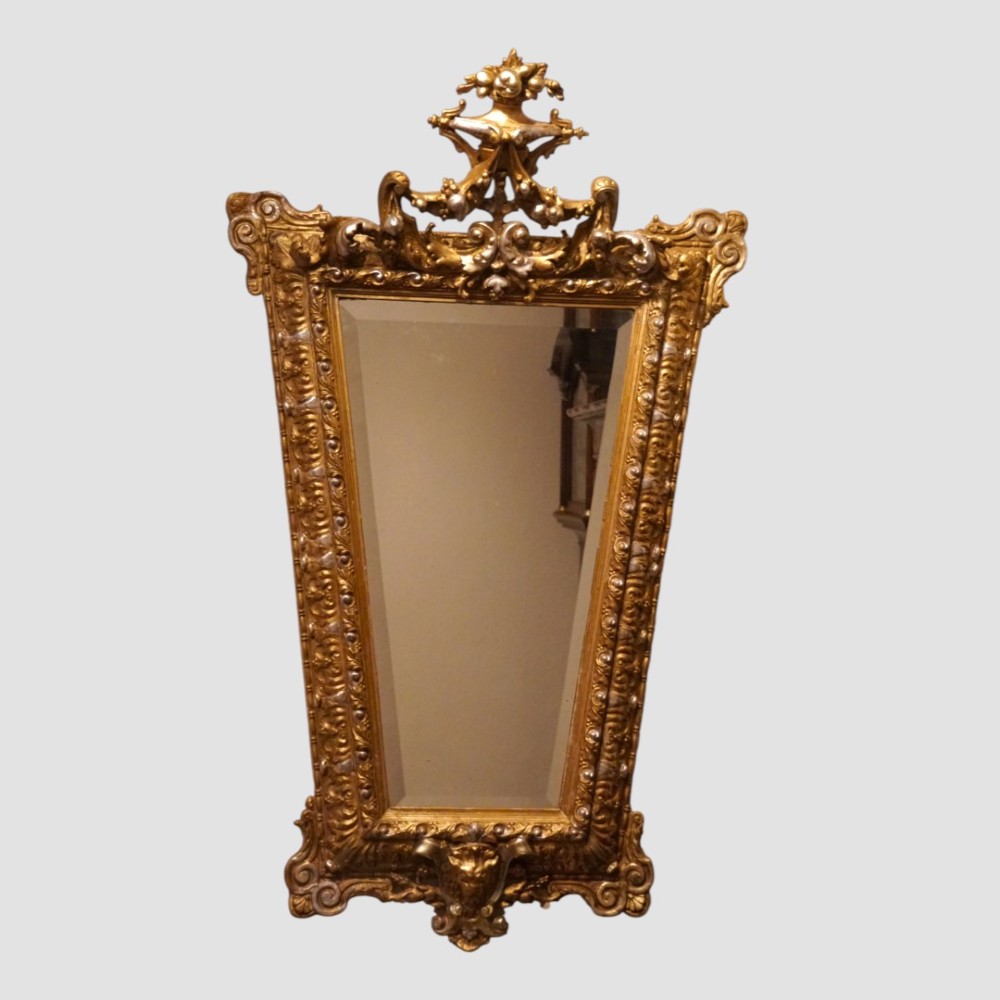 An antique trapezoidal wall mirror featuring an ornate giltwood frame with silver-gilt accents, a decorative carved crest, and a tapered profile.