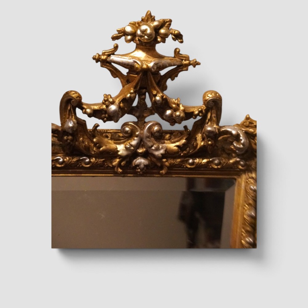 An antique trapezoidal wall mirror featuring an ornate giltwood frame with silver-gilt accents, a decorative carved crest, and a tapered profile. View III