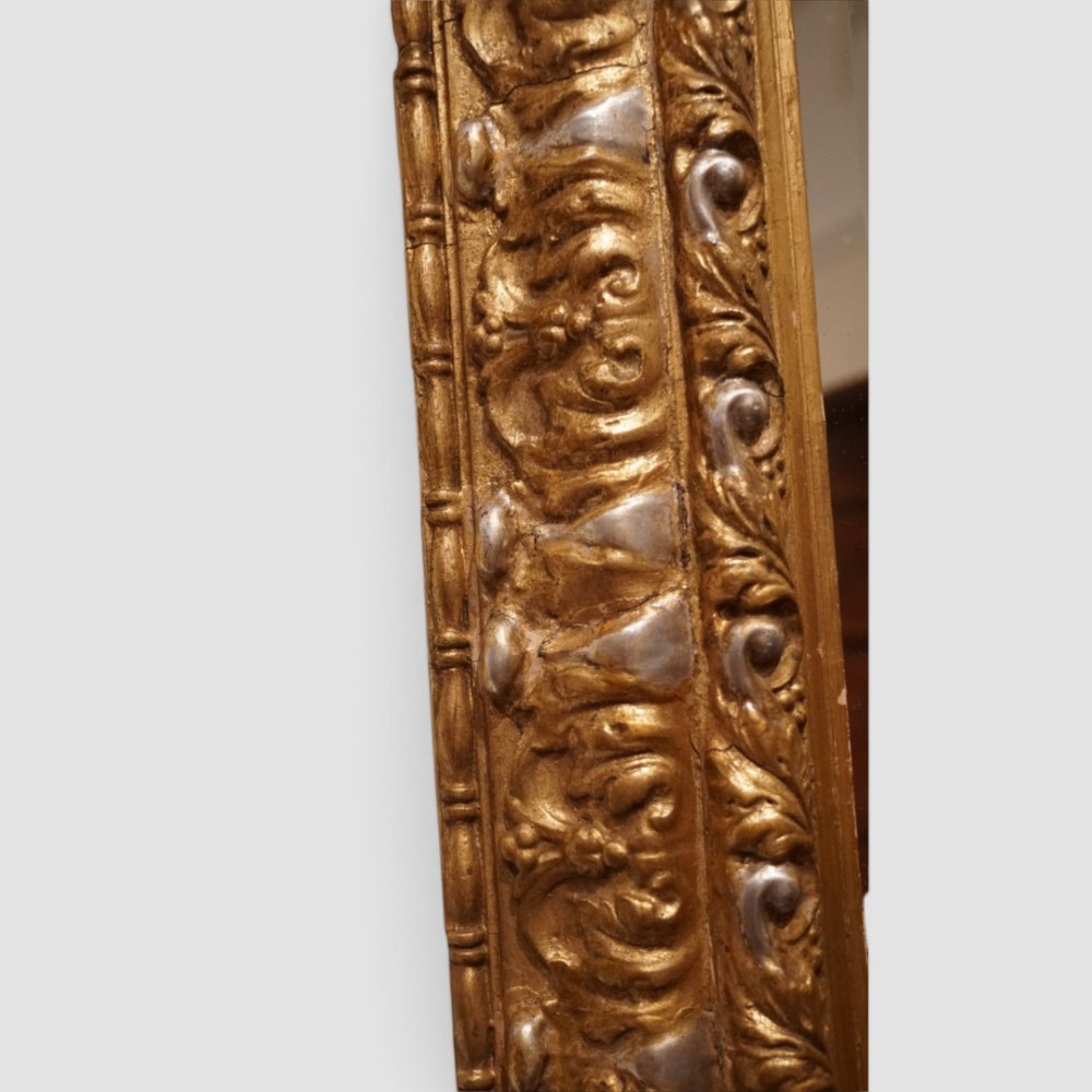 An antique trapezoidal wall mirror featuring an ornate giltwood frame with silver-gilt accents, a decorative carved crest, and a tapered profile. View V