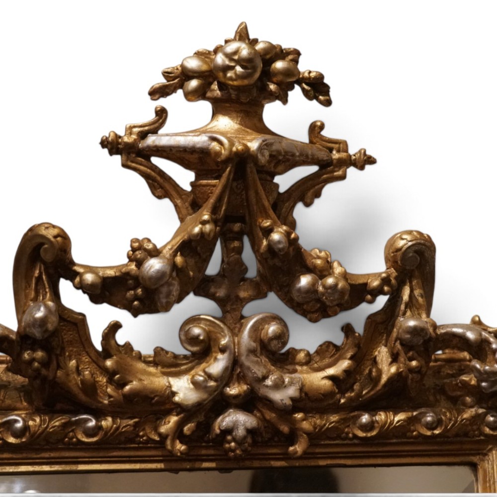 An antique trapezoidal wall mirror featuring an ornate giltwood frame with silver-gilt accents, a decorative carved crest, and a tapered profile. View VI
