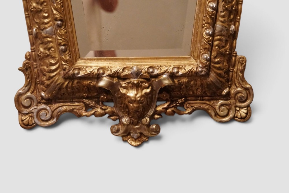 An antique trapezoidal wall mirror featuring an ornate giltwood frame with silver-gilt accents, a decorative carved crest, and a tapered profile. View VIII