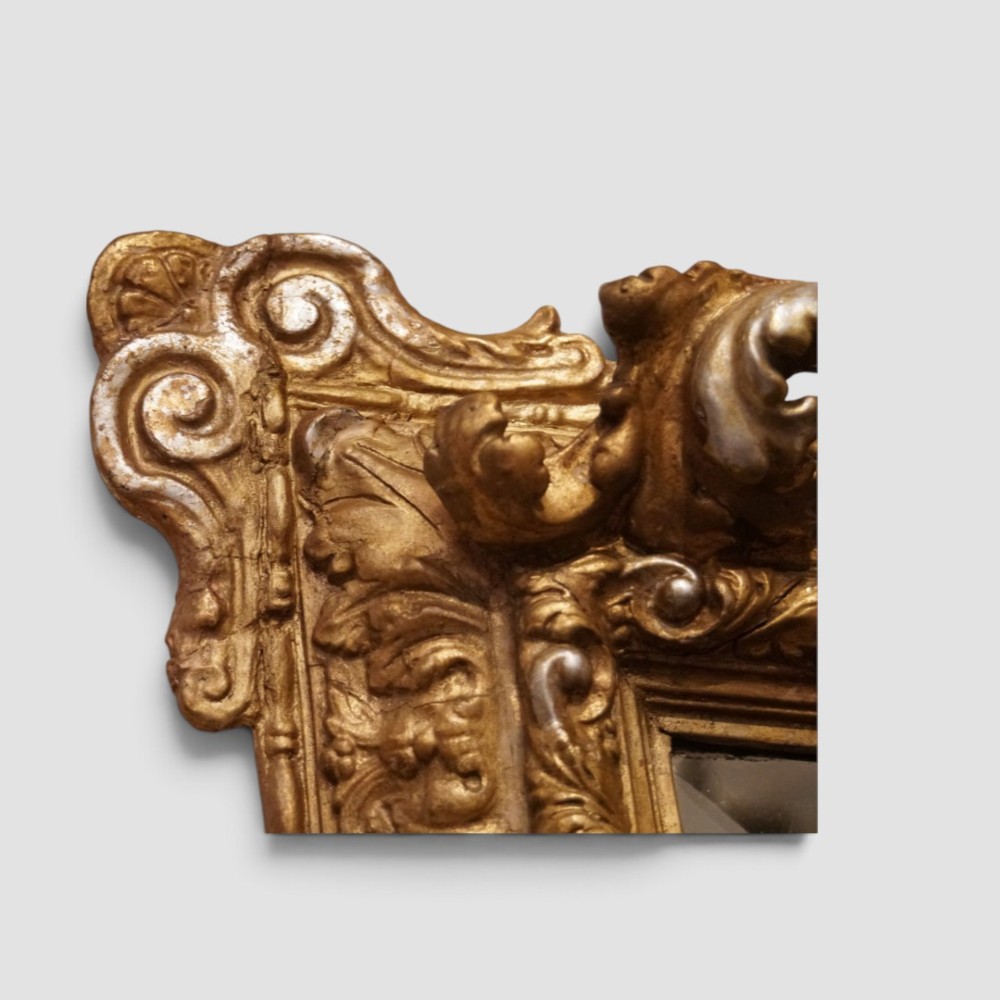 An antique trapezoidal wall mirror featuring an ornate giltwood frame with silver-gilt accents, a decorative carved crest, and a tapered profile. View IX
