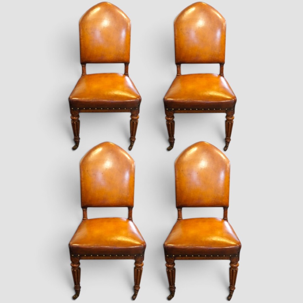 A set of four Victorian oak dining chairs featuring arched gothic-style backrests upholstered in warm brown leather with brass stud trim.