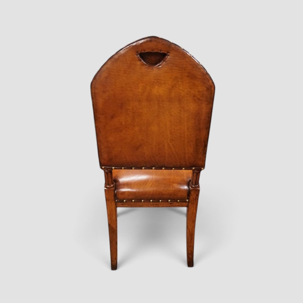 A set of four Victorian oak dining chairs featuring arched gothic-style backrests upholstered in warm brown leather with brass stud trim. View IV