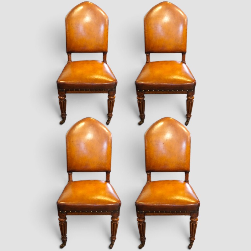A set of four Victorian oak dining chairs featuring arched gothic-style backrests upholstered in warm brown leather with brass stud trim. View V