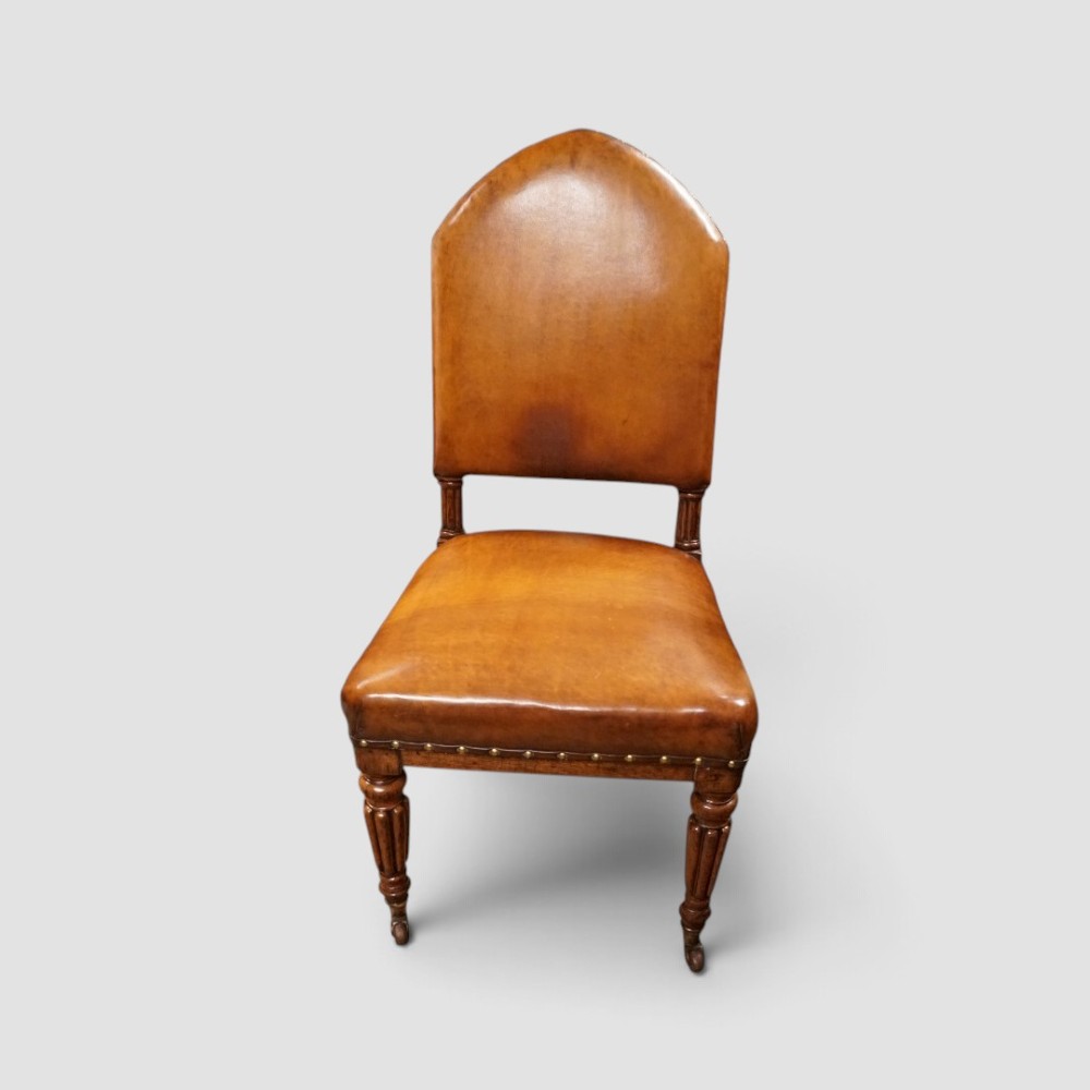 A set of four Victorian oak dining chairs featuring arched gothic-style backrests upholstered in warm brown leather with brass stud trim. View VII