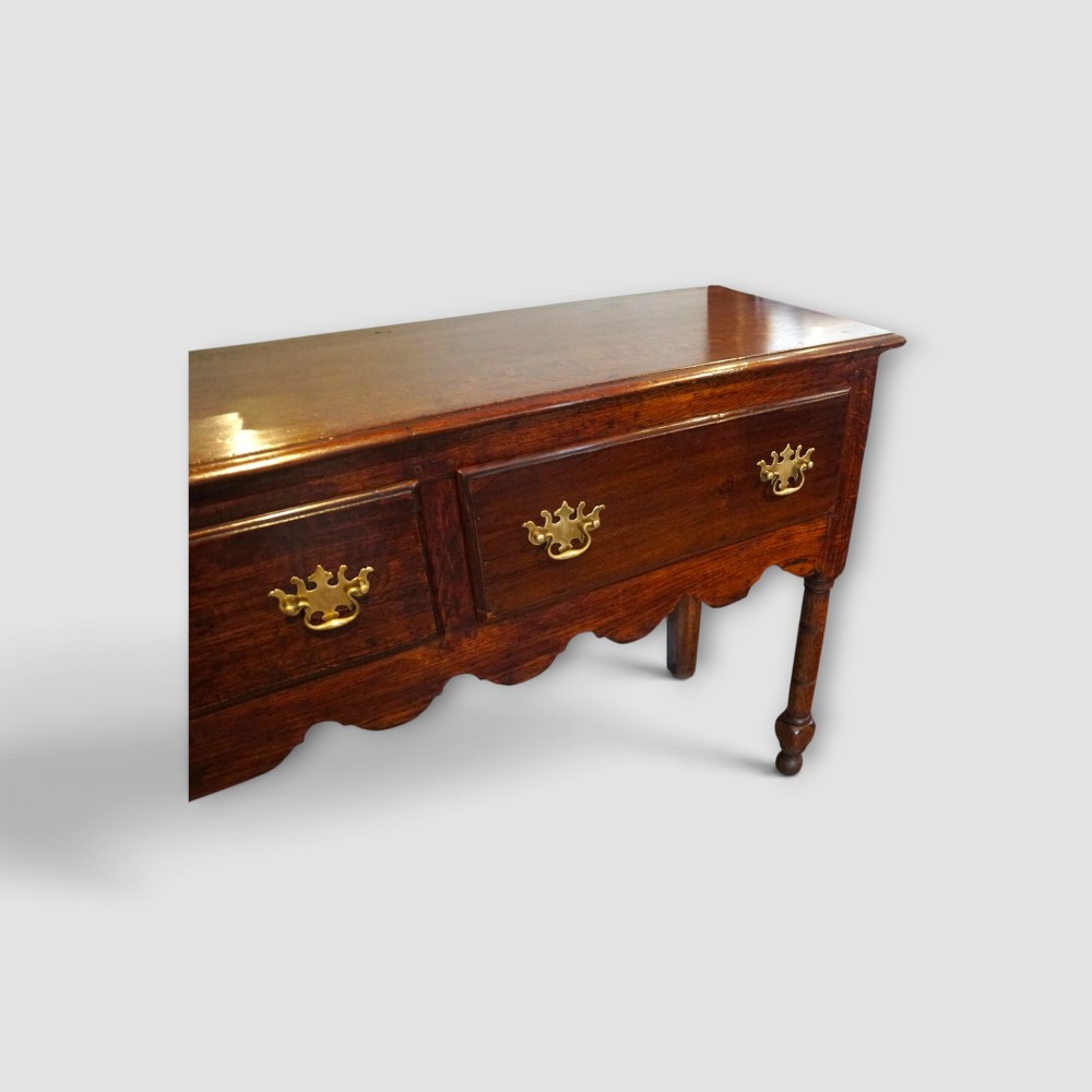An 18th-century George III dark oak dresser base featuring three drawers with brass handles and a scalloped apron on turned legs. View II