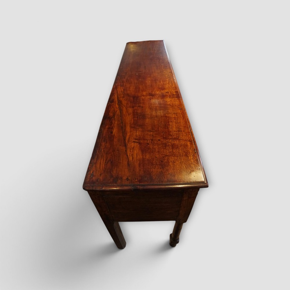 An 18th-century George III dark oak dresser base featuring three drawers with brass handles and a scalloped apron on turned legs. View X
