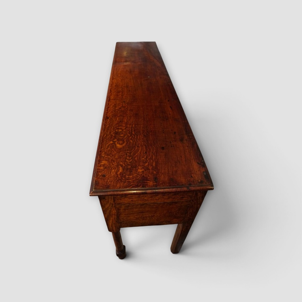 An 18th-century George III dark oak dresser base featuring three drawers with brass handles and a scalloped apron on turned legs. View XII