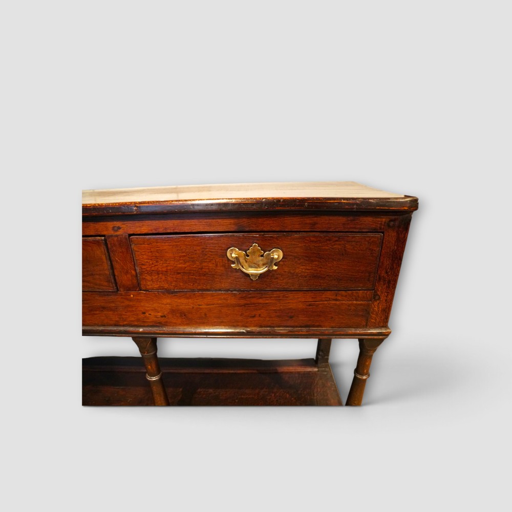 An antique dark oak dresser base featuring three frieze drawers with brass handles supported by turned legs and a low open potboard shelf. View VI