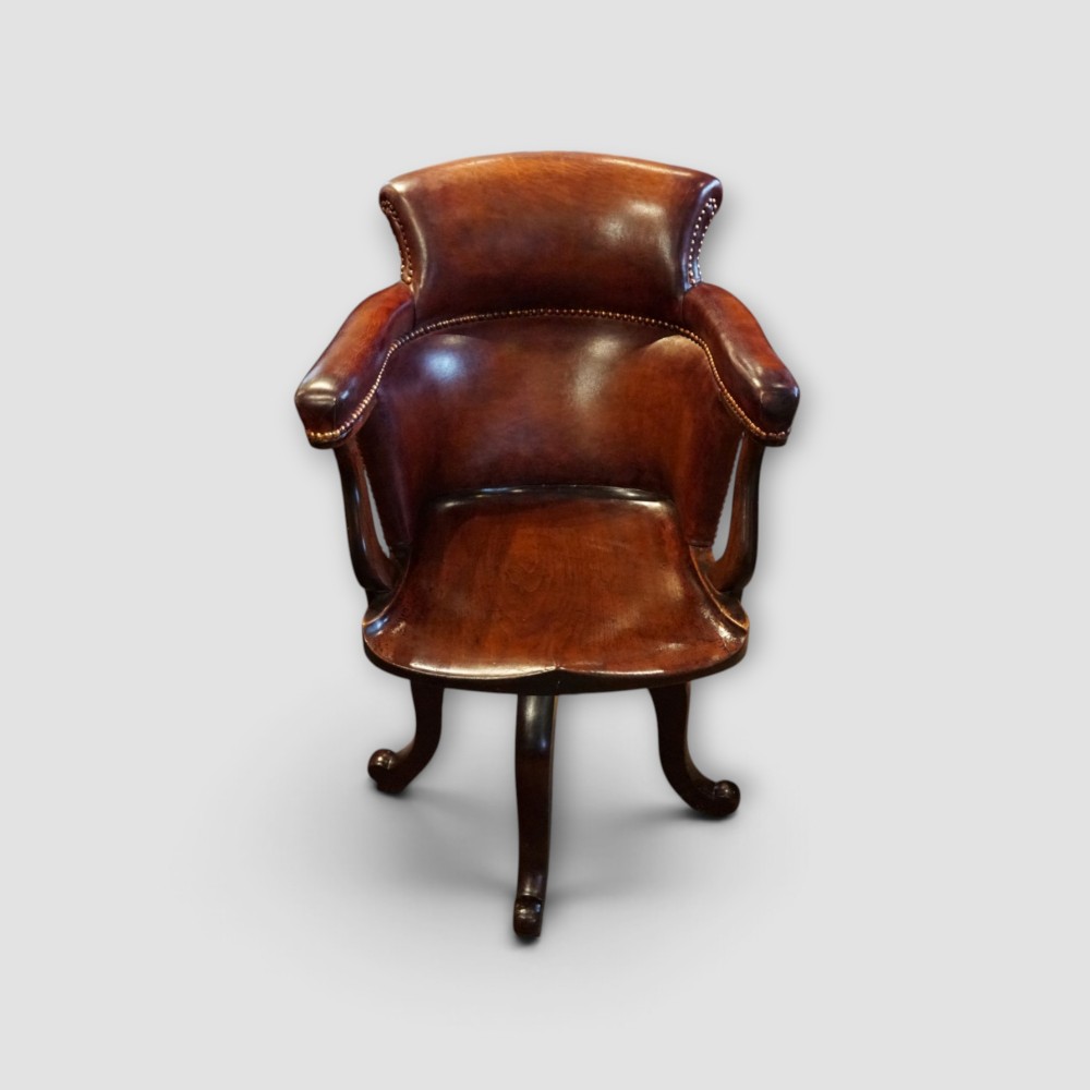 An Edwardian mahogany and chestnut brown leather swivel desk chair with a curved tub backrest, brass stud detail, and a carved four-star base.