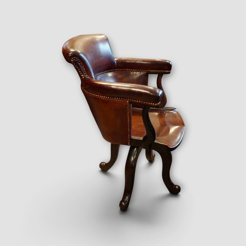 An Edwardian mahogany and chestnut brown leather swivel desk chair with a curved tub backrest, brass stud detail, and a carved four-star base. View VI