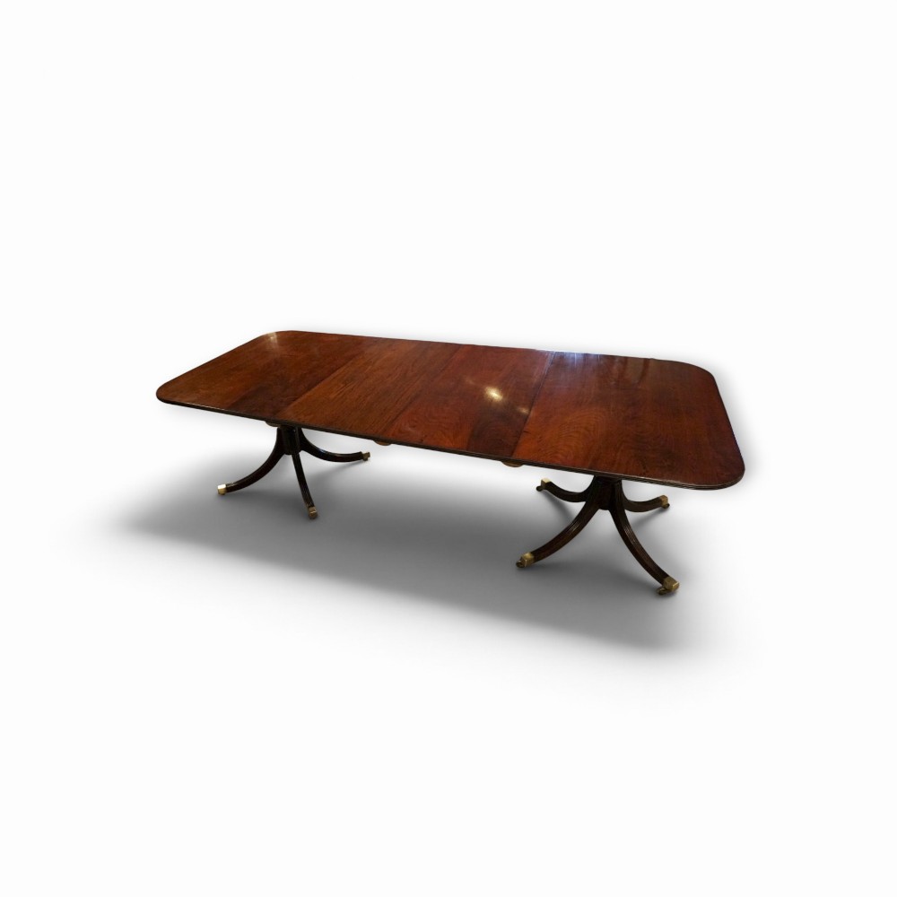 A Georgian style dark mahogany dining table featuring an extendable top, twin turned pedestal pillars, and splayed quadruped legs with brass toe caps.