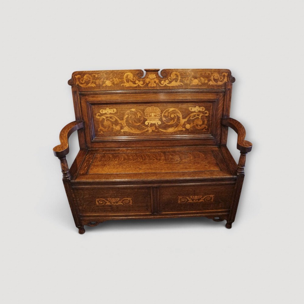 An Art Nouveau solid oak settle featuring decorative marquetry inlay across the backrest and lower front panels, curved armrests, and a lift-up storage seat.