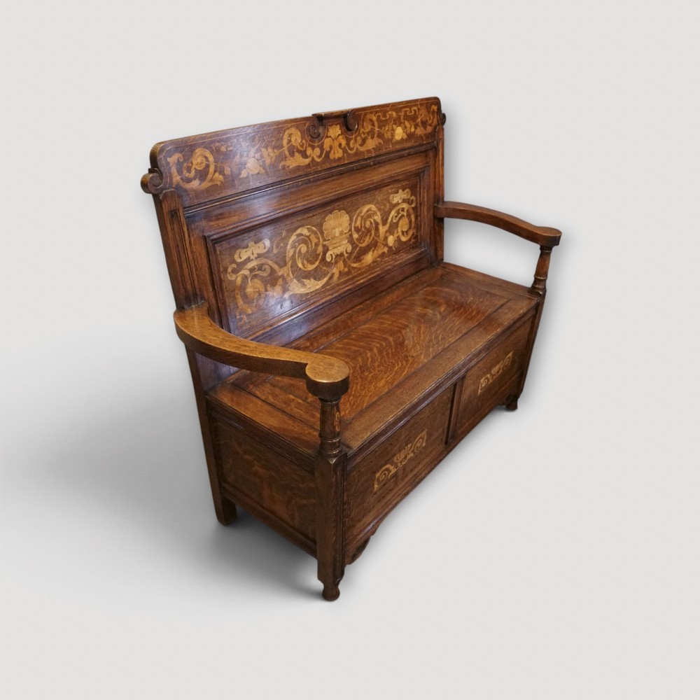 An Art Nouveau solid oak settle featuring decorative marquetry inlay across the backrest and lower front panels, curved armrests, and a lift-up storage seat. View II